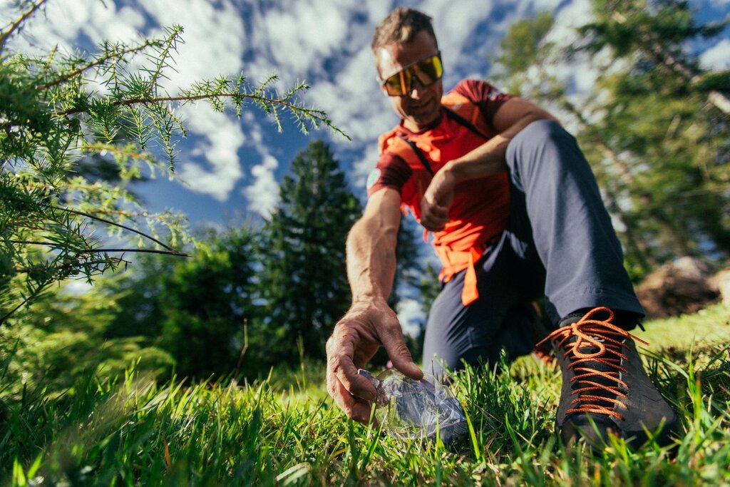 Un uomo raccoglie una bottiglietta di plastica accartocciata abbandonata lungo un sentiero in montagna