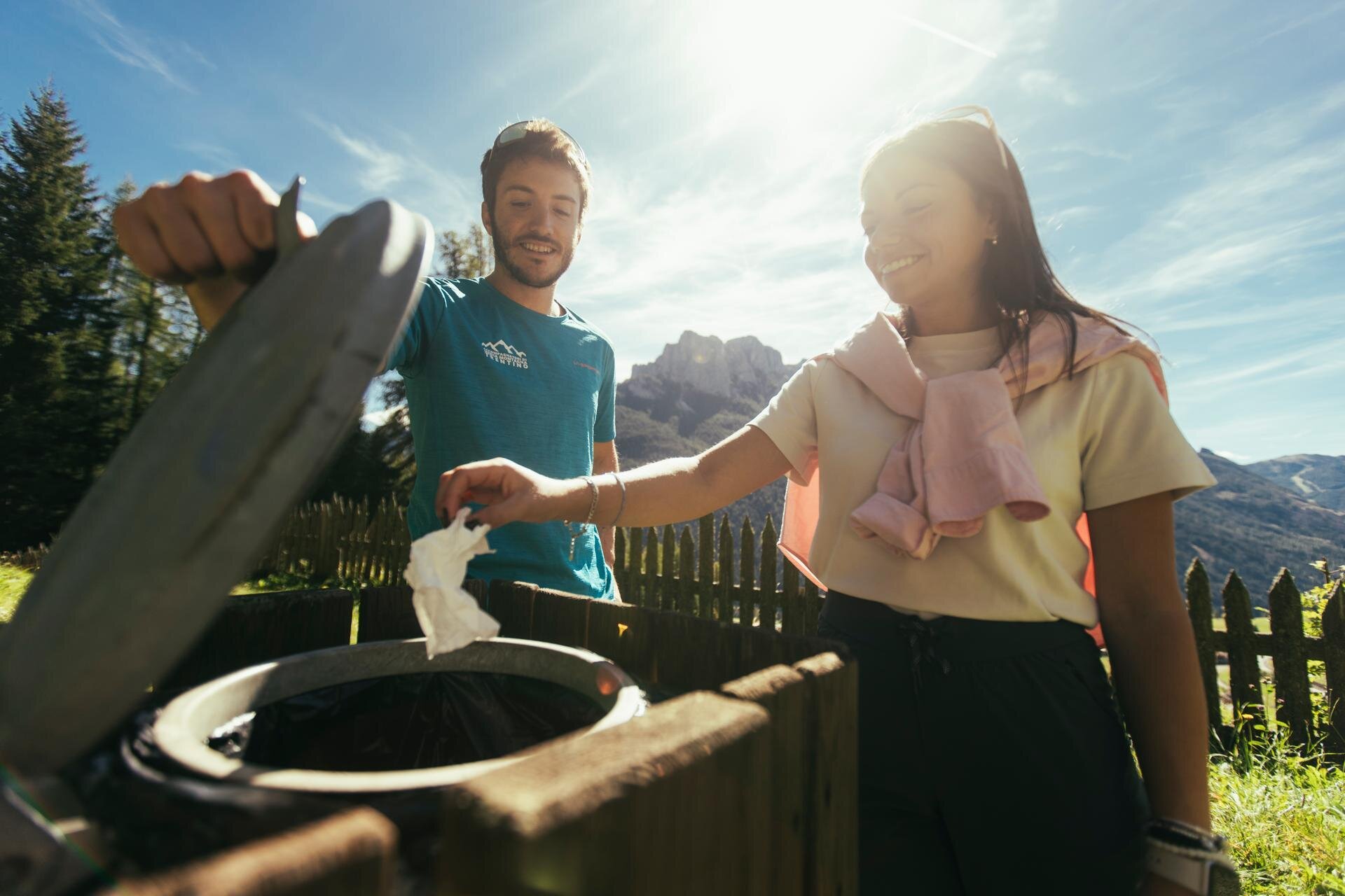 Un ragazzo apre un bidone della spazzatura mentre una ragazza butta un fazzoletto, sullo sfondo le Dolomiti di Fassa | © Archivio Immagini ApT Val di Fassa - Federico Modica