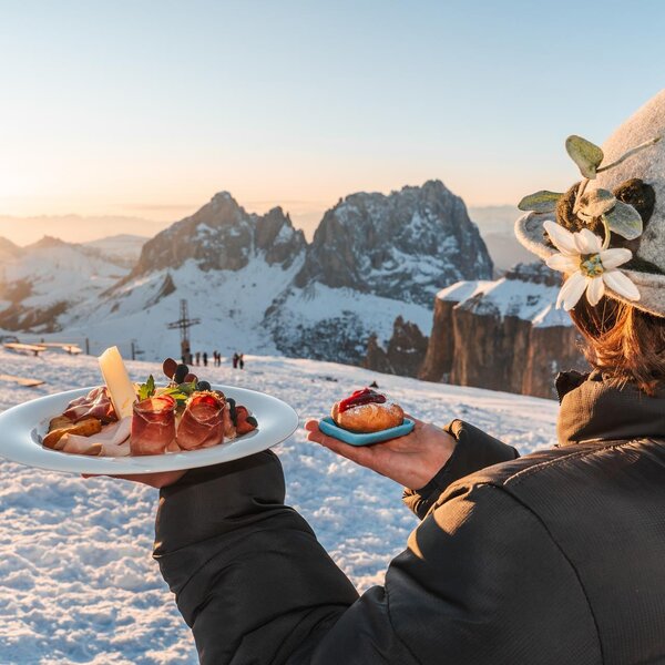 Ragazza con copricapo tipico regge piatti della tradizione al Sass Pordoi con vista sul tramonto sul Sassolungo | © Archivio Immagini ApT Val di Fassa - Patricia Ramirez