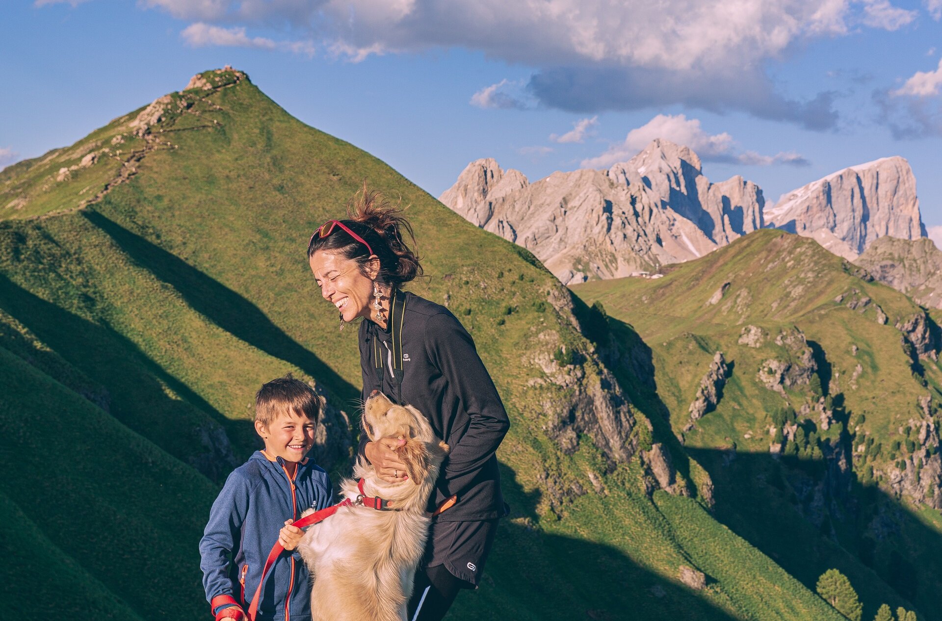 Donna e bambino sorridenti con cane in montagna verde e cielo sereno con ghiacciaio della Marmolada sullo sfondo | © Patricia Ramirez  - Archivio Immagini ApT Val di Fassa