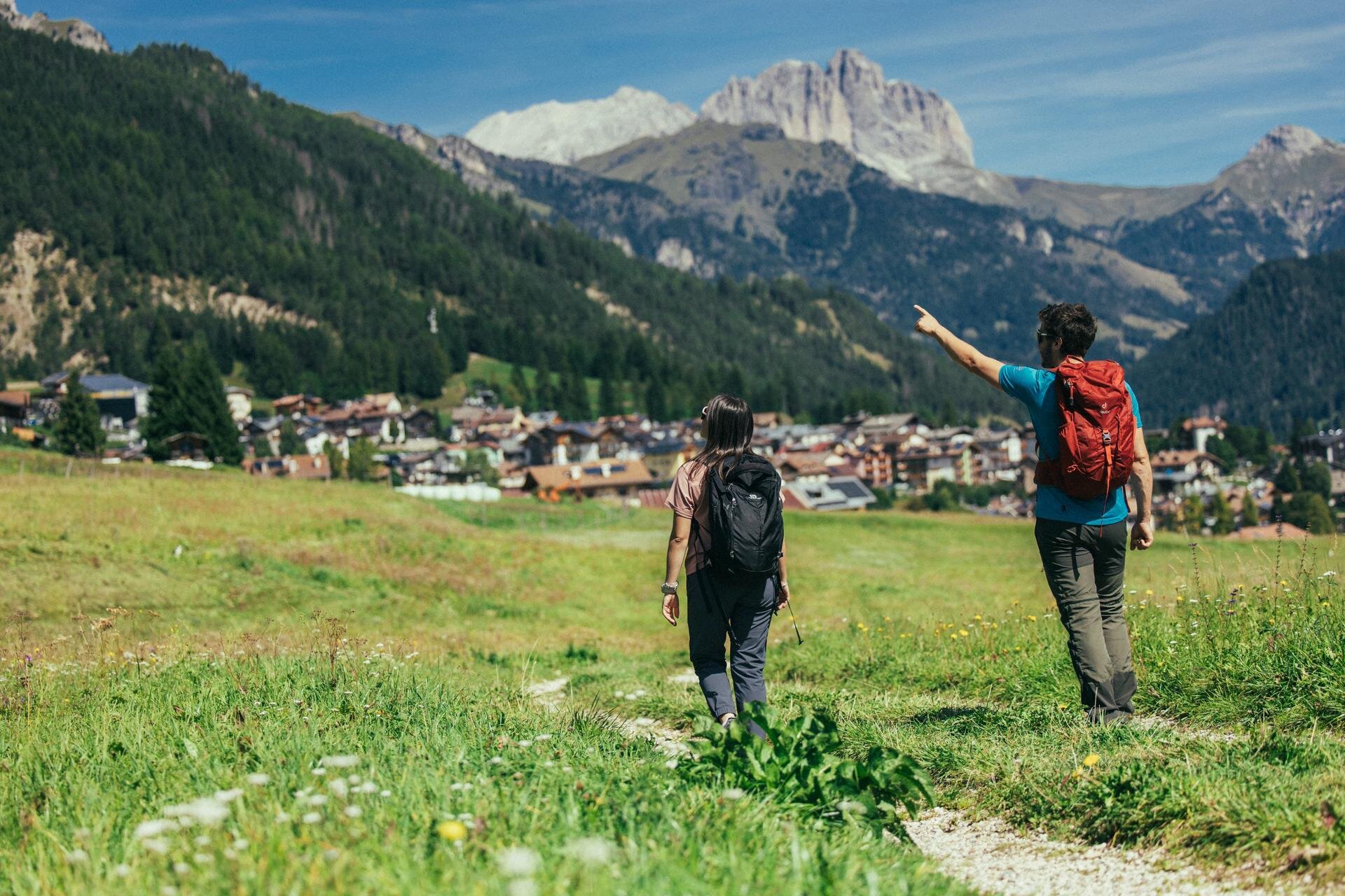 Coppia di spalle cammina in un prato estivo con vista sulle Dolomiti di Fassa | © Archivio Immagini ApT Val di Fassa - Federico Modica