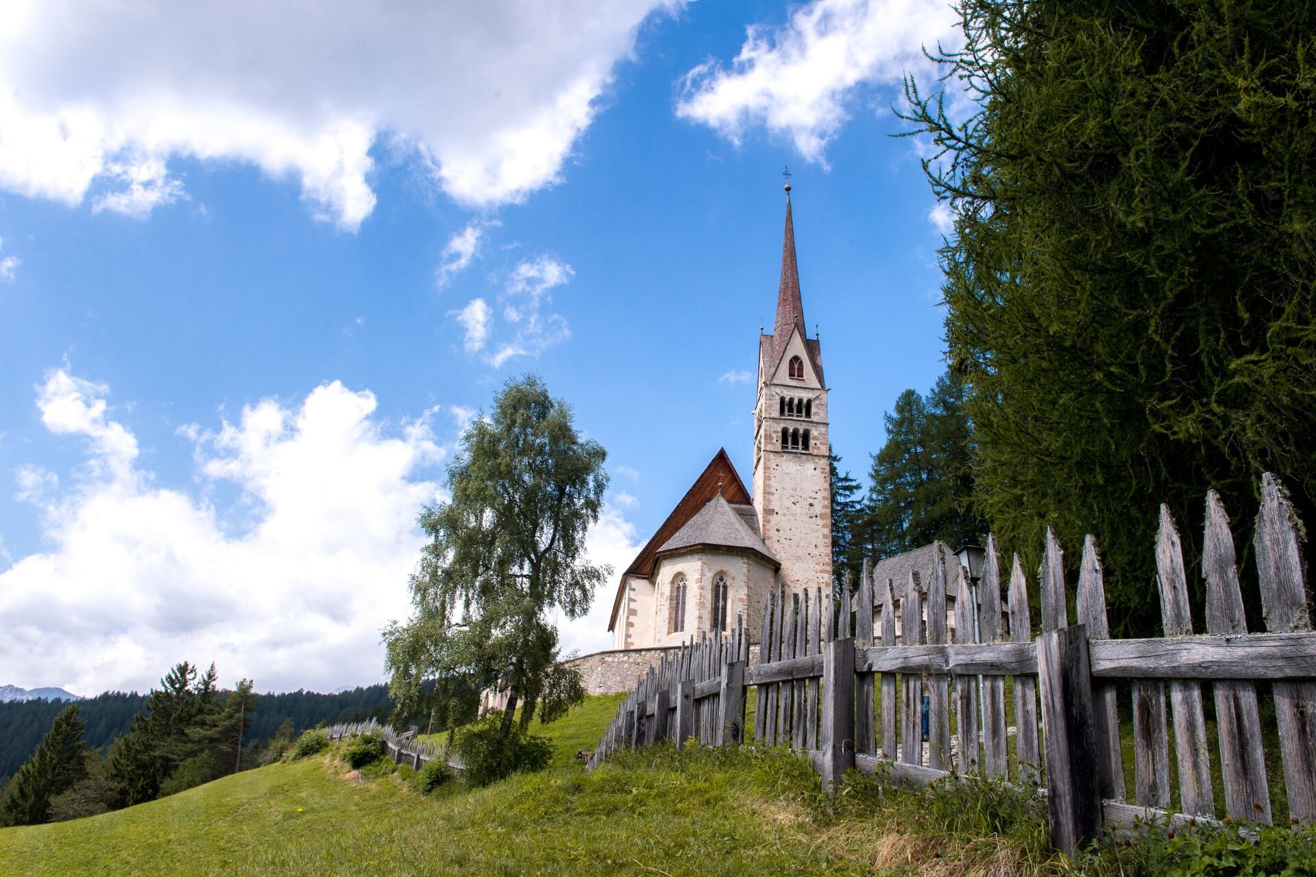 Visuale di una chiesa con campanile sul limitare del bosco, delimitata da una staccionata | © Archivio Immagini ApT Val di Fassa - Mattia Rizzi
